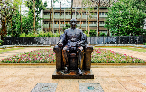 Bronze statue of Sun Yat-sen sitting in a chair with a flower bed in the background.