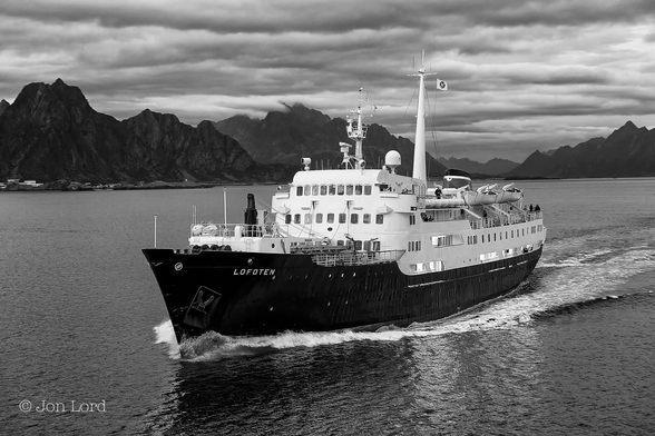 This is a black and white photo in landscape format of the historic Hurtigruten former ferry, MS Lofoten.
The lower two-thirds of the image is filled with calm, dark, sea water, with just a few small ripples disturbing the surface. In the centre of the frame is an oblique view of an old Norwegian ferry. The ship is underway from right to left, moving diagonally across the image with the bow and port (left) side of the ship in view, with her filling about half the length of the photo. The ships hull is black while the superstructure is white. The first third of the deck is taken with a small crane and forward cargo hatch. Moving aft is the superstructure with three rows of forward facing windows, the upper ones being the ships bridge. Aft are three decks, the lower one is partially open, the middle deck is enclosed, the upper deck, open with two large lifeboats suspended from davits. Above the wheelhouse is a collection of radio masts and radar antennas, in the centre of the ship is a tall signal mast with the shipping lines pennant flying from a halyard. A little further aft is a small funnel. A few of the ship's passengers can be seen on deck. In the distant background, perhaps ten km away, is a tall, dark and ragged range of mountains that stretch from the left to right, with a few scattered building in view on the shore. Above is a thin, near overcast sky.

