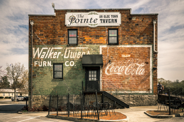 Historic two-story brick building with weathered signage showing "The Pointe: An Olde Town Tavern" at the top. The aged brick facade displays faded vintage advertisements including "Walker-Dunn Furn. Co." painted in white on a darker section and a classic Coca-Cola logo with "Enjoy" text on the right side. The building features four windows with dark frames, a black door with an overhead awning, and exterior stairs with metal railings leading to the entrance. A small fenced patio area with outdoor furniture is visible at ground level. The sky shows scattered clouds, and bare trees are visible in the background along with parked vehicles and neighboring buildings.