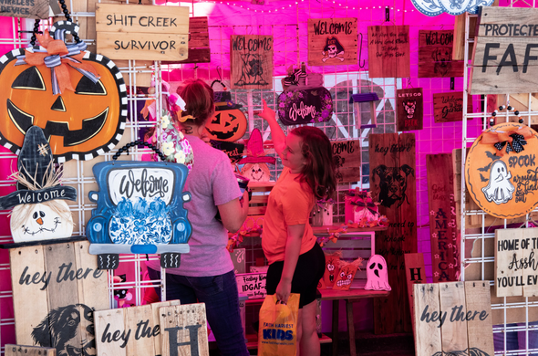 A photo of the inside of a traitor's tent during an autumn festival.  There are many items hanging inside and outside the stall.  Inside there is a young girl showing her mother a wooden sign with a depiction of a dog with the word "Welcome".