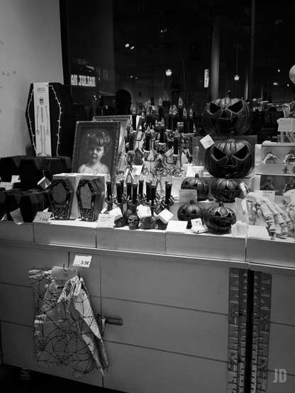 La imagen en blanco y negro muestra un expositor de tienda con artículos de decoración de Halloween.
Los elementos centrales son varias calabazas talladas con rostros terroríficos, réplicas de ataúdes (algunos con precios como 7,50€), y pequeños objetos como calaveras y velas oscuras. Un detalle notable es un retrato de un niño pequeño enmarcado, colocado en el centro, que contrasta con la temática macabra circundante.
Es una escena que evoca la estética de la "Noche de Brujas".