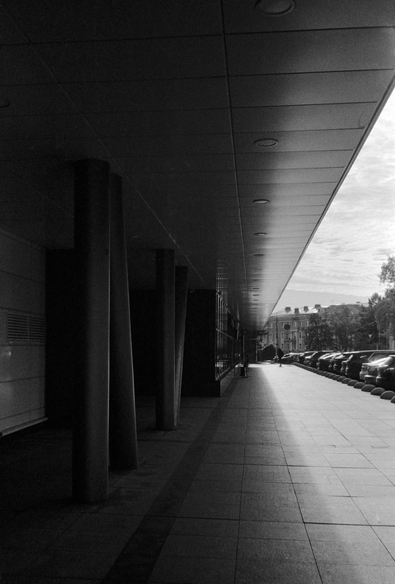 The black-and-white photo shows a ledge of a modern building that creates a tunnel effect. The ledge casts a shadow on the sidewalk, creating a clear boundary between light and shadow. Metal columns can be seen on the right. A silhouette of a person can be seen in the distance.