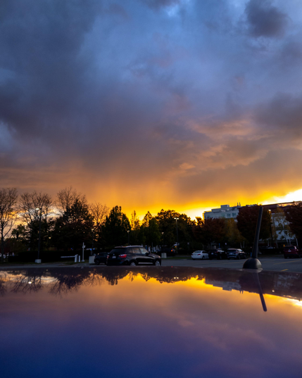 A yellow and orange sunset along a horizon bounded on the top by dramatic blue clouds and below by the sky’s reflection on the roof of a car.