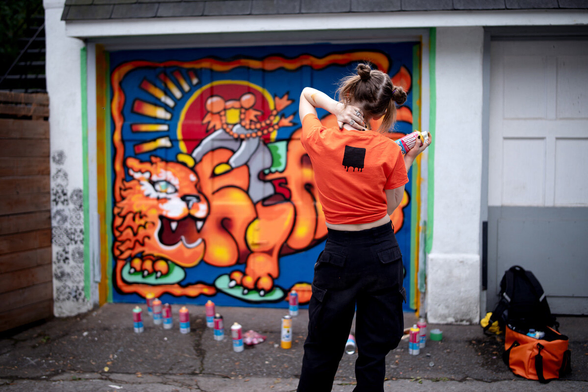 A spraycan female artist looking to her new wall, she is wearing orange and black. Her painting is representing her on a orange feline dragon, on a blue background.