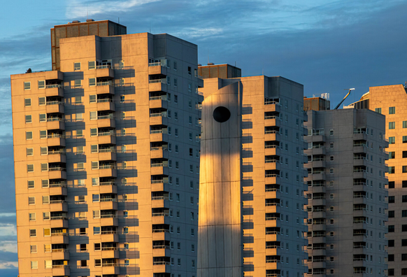 A row of identical, tall concrete apartment buildings with rows of windows and balconies stands in a row, yellowish from the evening sun against a blue sky (blue hour). The leftmost building is skewed (due to the photo) and deliberately not straightened. In the center, in front of the buildings, stands a tall concrete object in the same yellowish hue. It's a flat, wide bar of concrete elements with a slanted side on the left and a large, round hole at the top. The object appears almost as tall as the buildings. Because of the two opposing slanted lines, it seems to me as if the buildings have been shoved like cards in a hand.