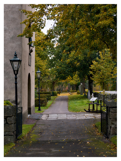 En stående bild på en gångväg av asfalt förbi ingången till en kyrka. Till vänster står en lampa på en stolpe vid en ingång i en stenmur. Bortanför kyrkan står många träd på kyrkogården i grönt och gult.