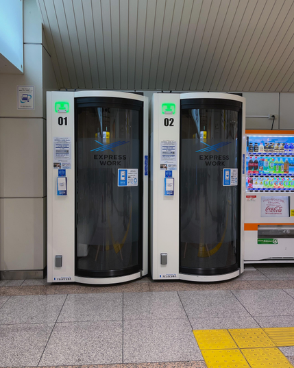 Un par de cabinas (parecen duchas, como un tubo con una parte algo circular donde va la puerta bastante opaca pero no al 100%) para aislarse y trabajar. Están junto a una máquina de vending. En el suelo asoman unas baldosas amarillas: en Japón se usan para que los invidentes puedan orientarse.