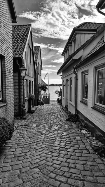Das Schwarzweißfoto zeigt eine Gasse mit Kopfsteinpflaster. Links und rechts davon stehen kleine gemauerte Häuser. Auf der linken Seite steht eine Straßenlaterne. Am Ende der Gasse befindet sich ein Fahnenmast mit einer schmalen Fahne, die im Wind weht. Danach ist ein See zu sehen, auf der gegenüberliegenden Uferseite sind Bäume und Buschwerk zu erkennen. Der Himmel ist durchzogen von Wolken.