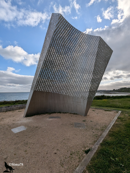 Monument en métal incliné, gravé de textes, face à la mer sous un ciel nuageux.
Slanted metal monument, engraved with texts, facing the sea under a cloudy sky.
Monumento de metal inclinado, grabado con textos, frente al mar bajo un cielo nublado.