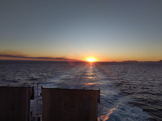 La mer au coucher du soleil, depuis le pont d'un cargo dont on aperçoit les deux pans métalliques (là  relevés à la verticale) qui servent à faire descendre les voitures. Le soleil est pikecdans l'axe du sillage, on n'en voit plus que la moitié derrière l'horizon. Sur la droite il y a une côte peu visible, sur la gauche une traînée de fumée.