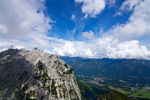 Im Vordergrund der Gipfel des Albspitzes. Blanker Fels mit einigen grünen Flecken. Darüber spannt sich ein locker bewölkter Himmel. Der Blick geht am Gipfel vorbei ins Tal, wo die Dörfer zwischen Wald und Wiesen liegen. 