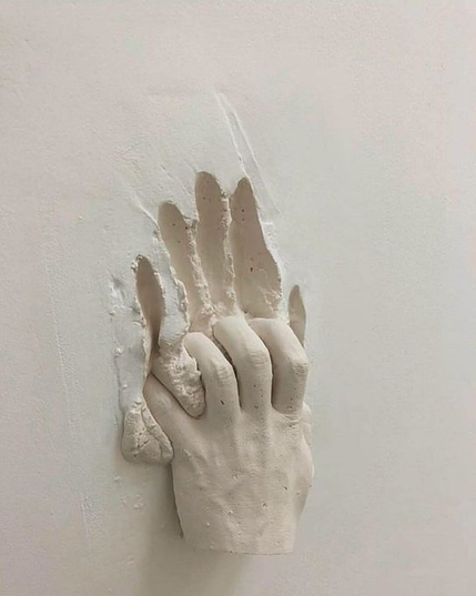 A photograph of a white plaster sculpture of two hands clasped tightly together, appearing to emerge from or sink into a plain white wall. One hand grips the other in a gesture that could be interpreted as either supportive or desperate, with the fingers digging into the plaster of the wall itself, leaving drag marks above the hands.