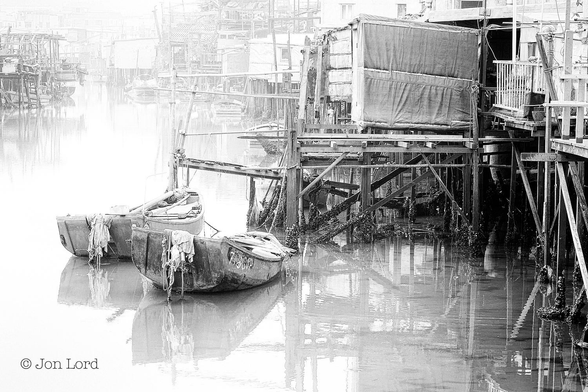 This is a black and white misty photo in landscape format of an old Chinese fishing village. Lantau (2014).
Stretching along the right margin and curving around to the left and into the distance is a long row of small houses or shacks (Pang Uks) all built on wooden stilts above a dead still and milky river. The shacks are randomly built, placed and in a state of disrepair, their reflections can be seen on the still water below. Many have small boats moored to the stilts. There is a thin mist about the village that becomes more obscured with increasing distance. Below and to the left of centre are two loosely moored dinghies, their sterns towards the camera and have oars left within the hull and a pile of what looks like rags dangled off the stern transom. In the distance and in the upper left corner is a misty view of further shacks on stilts. 
The village is Tai O, and active fishing community, located on the west end of Lantau Island, about 10 km from Hong Kong.