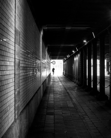The black-and-white photograph depicts a long, narrow pedestrian tunnel with tiled walls on the left and a row of columns on the right. The ground is paved with rectangular stones. A single person is walking away toward a bright light at the end of the tunnel, where daylight is visible.