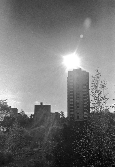 The black-and-white photo shows a high-rise building with a bright sun shining on its roof. In the foreground, you can see a park filled with sunlight and reflections.