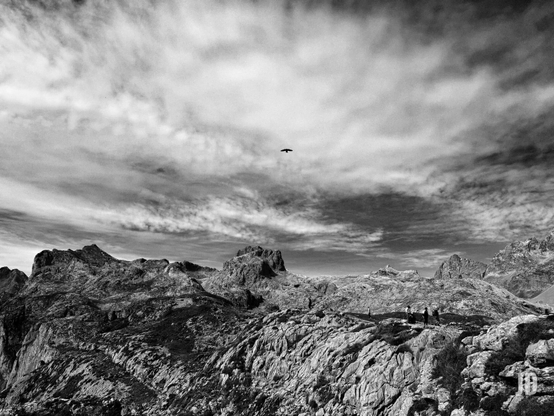 La imagen es una fotografía en blanco y negro que captura un paisaje de montaña rocosa y accidentada.
El terreno está compuesto por rocas irregulares y grandes formaciones montañosas. En el centro de la imagen, sobre el terreno elevado y rocoso, se distinguen a lo lejos tres figuras humanas pequeñas, que parecen ser excursionistas o montañeros, dando una idea de la escala del paisaje.
El cielo está ocupado por nubes dramáticas, algunas más densas y oscuras que otras, lo que añade profundidad y textura al ambiente. Justo encima del horizonte de las montañas, se ve la silueta de un pájaro solitario en vuelo.