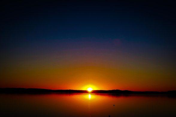 The image depicts a sunset over a body of water. The sky is a gradient of orange and red, transitioning from a brighter orange near the horizon to a darker red higher up. A bright, round sun is positioned just above the horizon line, appearing as a single point of intense light. Reflected in the water below is the sun, appearing as a similarly bright circular shape. In the distance, a dark silhouette of land is visible along the horizon line. There is no text present in the image.