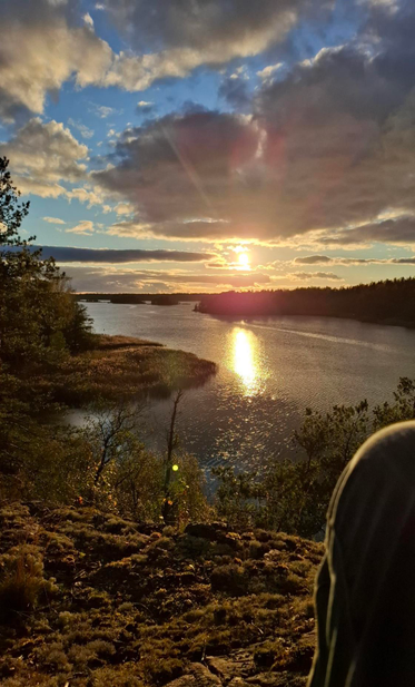 Soon-to-set autumn sun glinting on rippled water in a narrow inlet surrounded by reeds and trees, seen from a high rocky outcrop with a person's knee in the right foreground