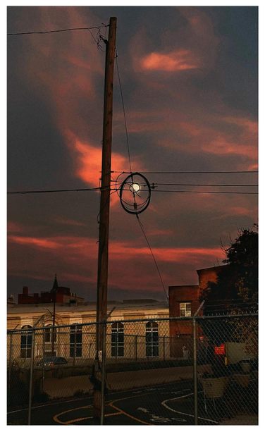 Photo of a city scene at sunset. Behind a chain link fence in the foreground, a road with yellow and white markings curves past some low buildings illuminated by the fading light. A tall wooden power pole rises into a darkening sky with luminous orange clouds. At the center of the image is the full moon, small in the sky, seen through some circular loops of cable attached to the power lines extending from the pole.