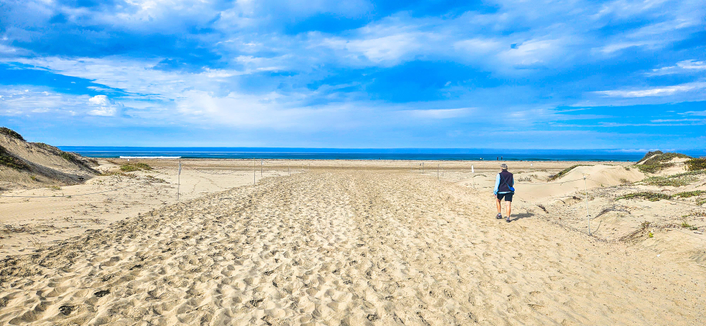 The image shows a wide expanse of sandy beach leading to a calm, blue ocean under a partially cloudy sky. A person stands near the water's edge, facing away from the camera, wearing dark shorts and a light-colored shirt. Footprints are visible in the sand leading towards the ocean. There are dunes on either side of the beach, partially covered in grass. The sky is blue with scattered white clouds, and the ocean stretches out to the horizon.