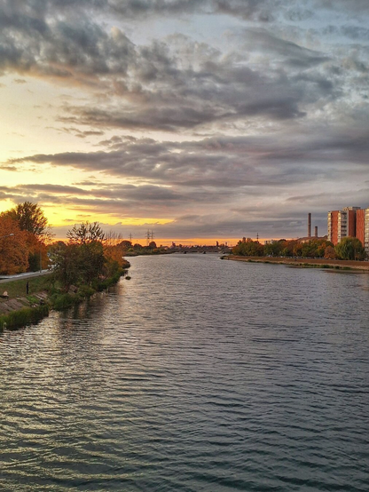 This is a landscape photo taken at sunset over a wide river. Dark, choppy water fills the foreground, reflecting the dramatic, cloudy sky with hues of gray, orange, and yellow. Autumn trees with orange and green leaves line both banks. On the far right, modern apartment buildings rise, contrasting with the natural scenery.