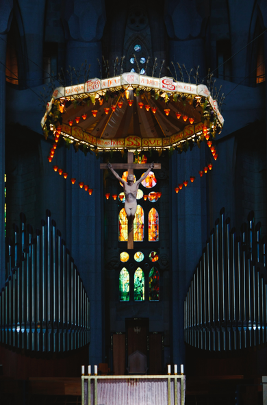 Colour photograph of Jesus on the Cross, surrounded by small candle-like lights and suspended above the organ in the Basilica de la Sagrada Família in Barcelona. The church interior lies largely in dim, blue shadow, contrasted by the light shining on the crucifix and through the stained glass windows in the background. The organ pipes are arranged symmetrically on either side of the crucifix and the windows. Although the picture shows only a very small portion of the church, the elaborate design and architecture gives an imposing sense of grandeur and the awe-inspiring scale of the basilica.