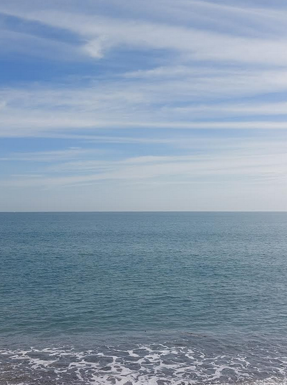 Photographie d'une mer calme avec un peu d'écume. Le ciel au dessus est bleu zébré de traits de nuages blancs.