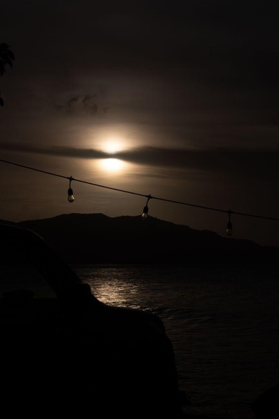 Silhouette of a Jeep and string lights against a hazy sunset over calm ocean water. The sun glows through thin clouds, reflecting softly on the sea with distant hills in the background.