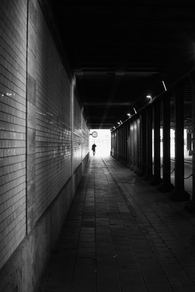 The black-and-white photograph depicts a long, narrow pedestrian tunnel with tiled walls on the left and a row of columns on the right. The ground is paved with rectangular stones. A single person is walking away toward a bright light at the end of the tunnel, where daylight is visible.