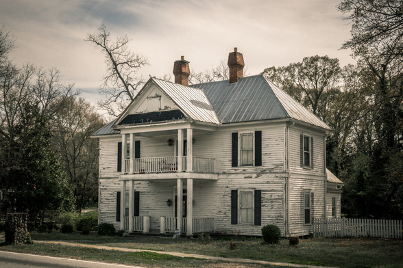 Weathered two-story white clapboard house with peeling paint and a gray metal roof featuring steep gables. The structure has two prominent brick chimneys and symmetrical black shutters flanking the windows. Double-stacked front porches with white railings span the facade, showing signs of age and wear. The house is surrounded by bare and partially leafed trees under an overcast sky, with a white picket fence visible on the right side and a paved road in the foreground. The overall scene conveys an abandoned or long-neglected rural property with faded Southern architectural charm.