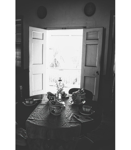 Black and white photo of a vintage dining room. Open double doors let in bright light, illuminating a table with ornate teacups and teapot, evoking nostalgia.