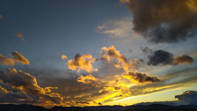 Evening photo of a partly cloudy sky. The clouds are lit yellow by the sun that is already behind the mountains on the horizon with some contrasting highlights and dark shadows.