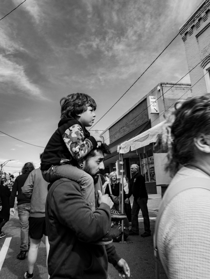 A black and white profile photo of a little boy on his father's shoulders taken at a street festival.