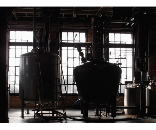 Dimly lit industrial scene with large brewing tanks and equipment silhouetted against bright windows. The setting feels utilitarian and atmospheric.