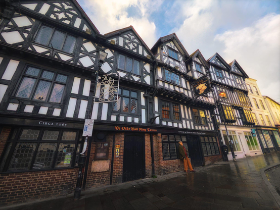 A striking row of historic timber-framed buildings in Ludlow displays the characteristic architecture of medieval England, with Ye Olde Bull Ring Tavern prominently featured at ground level, its brick frontage bearing the establishment's name in golden lettering beneath storeys of intricately patterned dark wooden beams and cream-coloured infill panels that rise to steeply pitched gable roofs. The building, dated circa 1365 as indicated on its left side, showcases multiple mullioned casement windows with leaded panes that reflect the afternoon light, whilst decorative wrought-iron signs hang from the facade, including a stylised tree motif above the tavern entrance. To the right, neighbouring buildings continue the architectural tradition with their own geometric beam patterns and stepped gables, their pale stone construction providing contrast, and the wet paving stones below suggest recent rainfall, all beneath a partly cloudy sky that bathes this quintessentially English historic streetscape in soft natural light, capturing the enduring charm and historical significance of this centuries-old market town building.