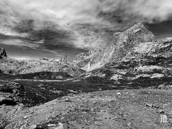 La imagen es una fotografía en blanco y negro que muestra un paisaje montañoso y accidentado.
En el primer plano hay un terreno rocoso y árido, con grava y algunas rocas más grandes. La vista se extiende hacia un valle o paso de montaña.
En el fondo, las montañas son escarpadas y prominentes, con un pico rocoso y masivo dominando el lado derecho. Las laderas de las montañas son muy rocosas y con mucha textura.
El cielo está cubierto de nubes dramáticas y con mucha textura, lo que contribuye a la atmósfera intensa de la fotografía en blanco y negro. La luz y el contraste resaltan las texturas del terreno y las montañas.