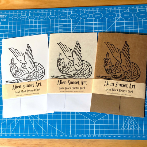 a set of three cards laid out on a blue cutting mat. a brown Kraft card, a plain white card, and a cream parchment pattered card. each card has a yellow parchment strip of paper wrapped around it that says "Alien Sunset Art" Hand Block Printed Card.

the block print on the cards is of  a a dragon standing it’s own tail like a platform spouting fire straight up with unfurled feathery wings