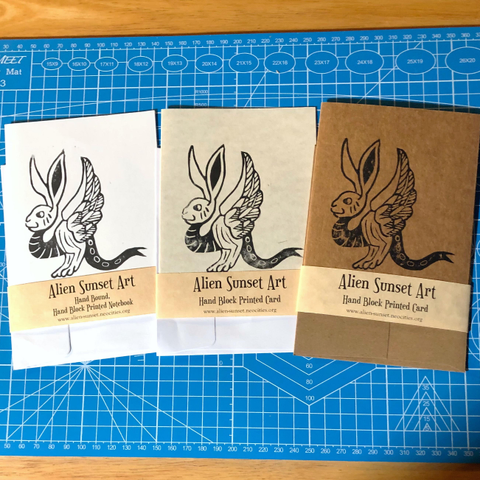 a set of three cards laid out on a blue cutting mat. a brown Kraft card, a plain white card, and a cream parchment pattered card. each card has a yellow parchment strip of paper wrapped around it that says "Alien Sunset Art" Hand Block Printed Card.

the block print on the cards is of  a creature with a bunny's head, upraised birds wings and forked lizards tail