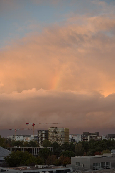 Paysage urbain au coucher du soleil. Nuages denses à la teinte très orangée. Au centre, on distingue un bout d'arc-en-ciel, quasiment noyé dans la lumière chaude.