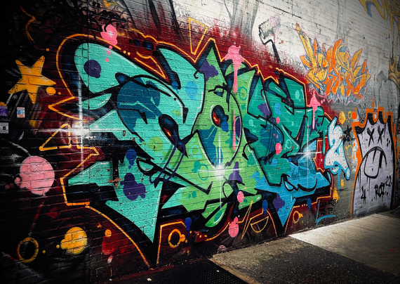 A color photograph in landscape orientation of a vibrant and dynamic graffiti art mural covering a weathered brick wall on a city street. The primary piece of graffiti is a complex "wildstyle" mural dominated by shades of teal, lime green, and dark blue, with bold black outlines and highlights of bright yellow-orange and hot pink dots and accents. The lettering is highly stylized and interlocking, and its energetic, angular shapes give the composition a strong sense of movement. The mural extends across the center and left of the frame, with smaller, older, and less defined tags and pieces visible on the right. In the far right background, a faded white and gray character-style piece is partially visible, offering a stark contrast to the main vibrant work. The photo is taken at street level, looking slightly up and along the wall, which recedes into the distance on the right.