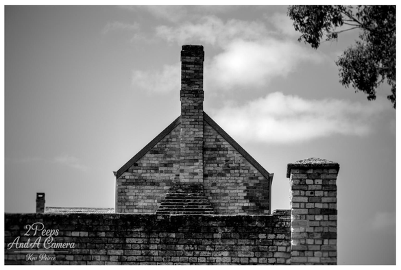 Black and white photograph of the gabled brick roofline and tall central chimney of a historic building at Port Arthur, Tasmania.

A lower, shorter brick column stands on the right, and a smaller chimney is visible on the far left. The structures are set against a bright, cloudy sky with dark eucalyptus leaves fringing the top right corner.