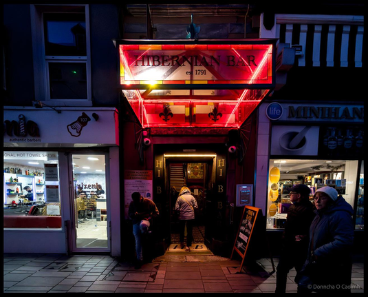 The illuminated red and gold Art Deco-style entrance to the Hibernian Bar (Hi-B) established 1791 on Oliver Plunkett Street in Cork at night, with people queuing to enter, flanked by authentic barbers shop on the left and Minihan pharmacy on the right, with the pub's distinctive vintage signage glowing against the dark evening.