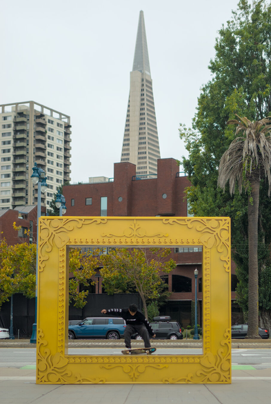 A skater framed by a large art piece that looks like a gilded frame for a painting, with the Transamerica pyramid in the background