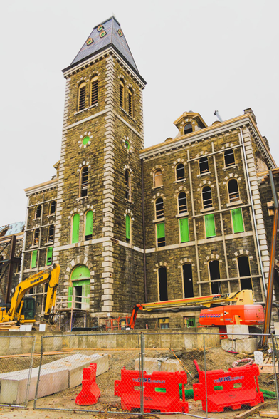 Sone building wit tall tower has windows filled up with queen panels with fences, orange barricades and yellow excavators in front