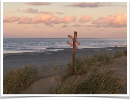 Clouds bathed in apricot light by the setting sun, above the blue north sea - that is reflecting the light of the sky. We look to the north-east from above a dune, down to the dark beach - with two people walking.
In the foreground marram gass grows on a dune, that is marked by a faded orange wooden post - as a place with a crossing. 