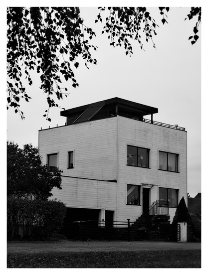 En stående svartvit bild på ett fyrkantigt vitt trähus med fyrkantiga fönster och en svart fyrkantig vind ovanpå taket. Grenar med löv hänger ner i förgrunden.