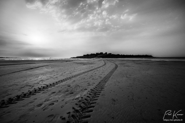 Photographie monochrome en noir et blanc présentant un paysage de bord de mer en fin de journée, avec notamment dans la moitié inférieure du cadrage une large plage de sable fin où on peut voir les traces de pas de promeneurs, mais aussi les traces de pneus d'un tracteur agricole.
Les promeneurs et le paysan reviennent d'une île maintenant cernée par la marée montante qui bientôt effacera leurs traces sur le sable.
Au-dessus de l'île, le ciel est partiellement nuageux.