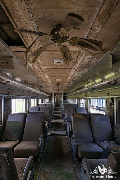 The interior of an abandoned train car with rows of worn seats, peeling ceiling paint, and a broken ceiling fan.