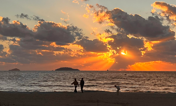 A sunset over the ocean, with vibrant orange and yellow hues illuminating the clouds. Silhouetted figures walk along the beach, with two people standing and a child crouching down behind them as if looking at the gray sand beneath her feet. Small islands are visible in the background. (Kkotji Beach, Chungcheongnam Province, S. Korea)