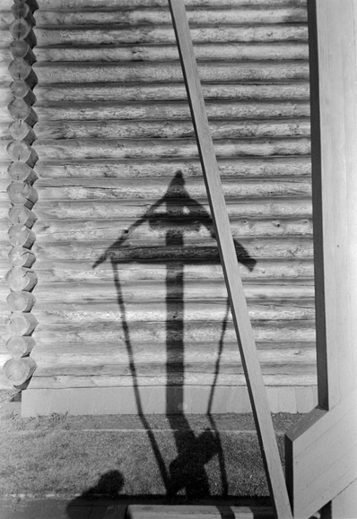 In this black and white photograph, a wooden cross casts a shadow on the wall of a wooden church.